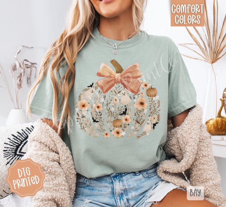 Pu&ograve; includere: T-shirt verde chiaro con un design a zucca pieno di fiori, pipistrelli e fantasmi. La zucca ha un grande fiocco color pesca e un gambo marrone. Le parole "Comfort Colors" sono stampate in alto a destra.