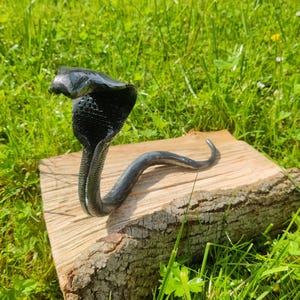 Könnte beinhalten: Eine handgefertigte Metall-Kobra-Skulptur mit aufgeweitetem Kopf und strukturierter Oberfläche. Die Schlange ist dunkelgrau und sitzt auf einem natürlichen Holzblock, vor grünem Gras.