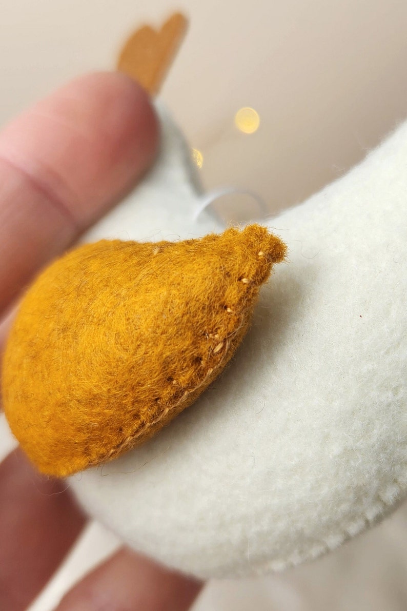 May include: A white felt ornament shaped like a bird with a yellow felt beak. The ornament is hand-stitched and has a rustic charm.
