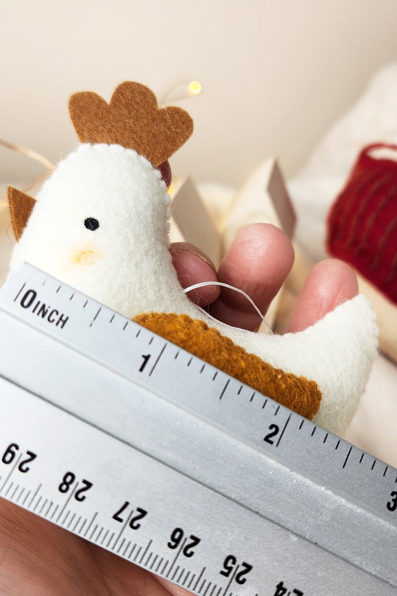 May include: A white felt chicken ornament with brown felt accents, measuring approximately 2.5 cm in length. The chicken is being held by a hand and a ruler is visible in the background.