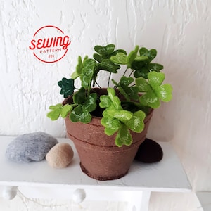 May include: A felt craft project of a potted plant with green four-leaf clover leaves in a brown pot. The pot is sitting on a white shelf with three gray felt rocks.