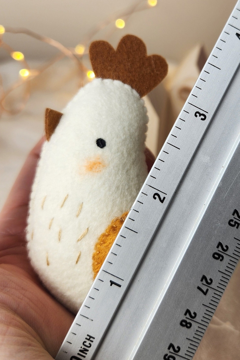 May include: A white felt chicken with a brown felt comb and wattle. The chicken is approximately 2.5 cm tall and is sitting on a ruler.