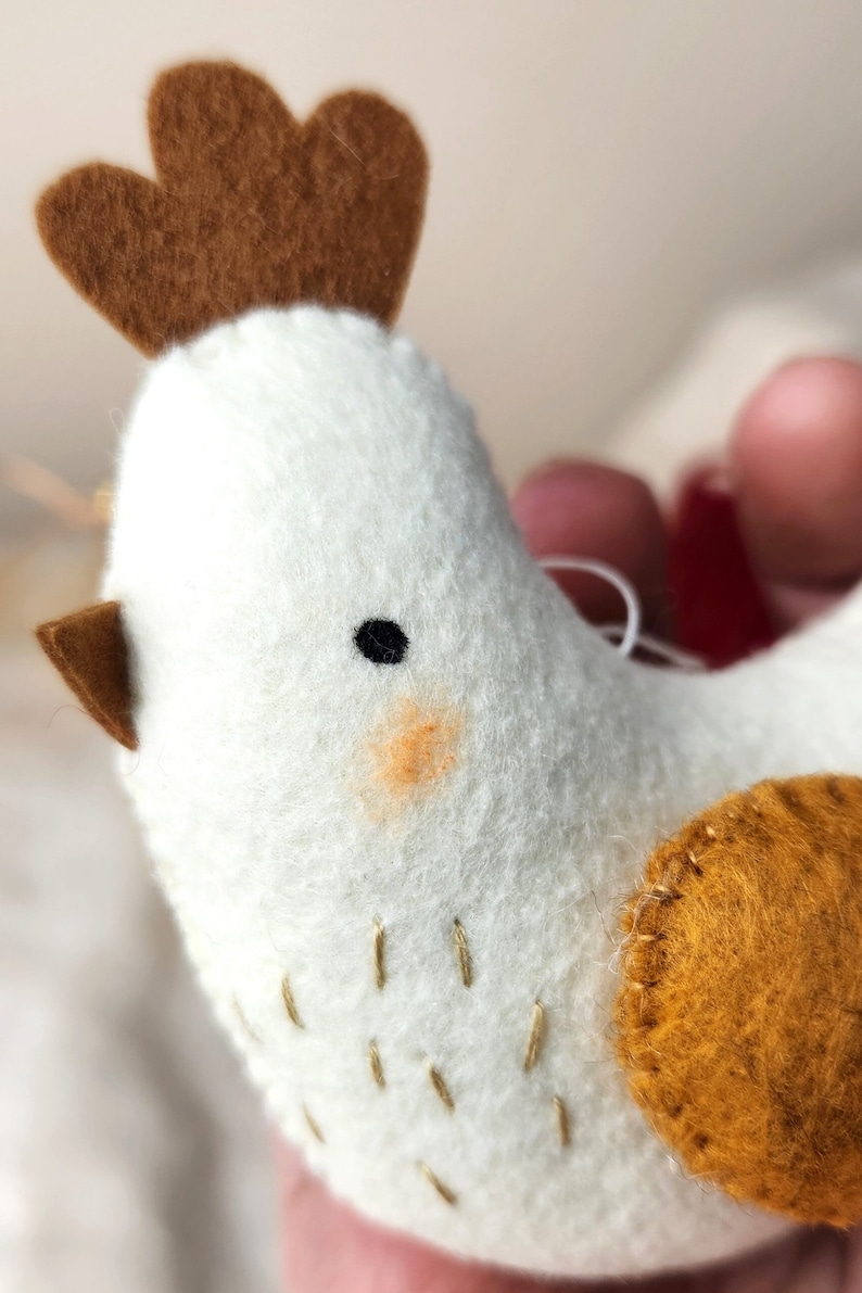 May include: A white felt chicken ornament with brown felt accents. The chicken has a brown felt comb, a brown felt beak, and a black eye. The chicken also has a small orange felt patch on its cheek. The chicken is decorated with gold stitching.