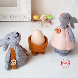 Puede incluir: Tres adornos de Pascua de conejo de fieltro, uno sosteniendo un porta huevos marrón con un huevo blanco dentro. Los conejos son grises y tienen ojos de botón marrones. Un conejo lleva un vestido rosa con un botón marrón. El texto "sewing pattern in" es visible en la esquina inferior derecha.