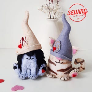 May include: Two felt cat and dog gnomes with pointed hats. The dog gnome is grey with black hair and a beige hat. The cat gnome is beige with brown stripes and a grey hat. Both have heart accents and are handmade.