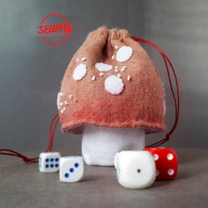 May include: A mushroom-shaped dice bag made of brown felt with white spots and a red drawstring. The bag sits on a white base, with four dice in front. The dice are white and red with blue and white dots. The text "sewing" is in a red circle.