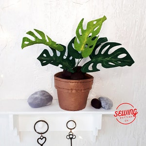 May include: A felt monstera plant in a brown pot with grey stones on a white shelf. The plant has green leaves with holes in them. The pot is sitting on a white shelf with a key holder underneath. The text "sewing pattern en" is in a red circle on the right side of the image.