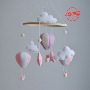 May include: A pink and white felt baby mobile with hot air balloons, clouds, and a star. The mobile is hanging from a wooden ring.
