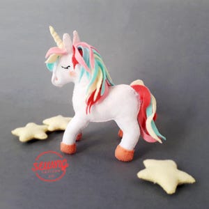 May include: A white felt unicorn with a multicolored mane and tail, a golden horn, and peach-colored hooves. The unicorn is surrounded by three yellow star-shaped felt decorations. The image has a soft, handmade aesthetic.