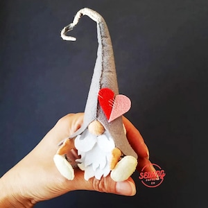 May include: A gray felt gnome with a long, curled hat and a white beard. The gnome is wearing a red and pink felt heart on its hat.