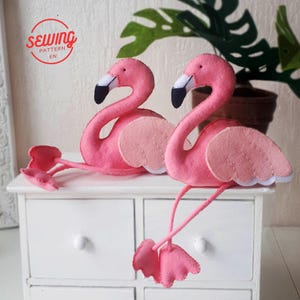 May include: Two pink felt flamingo ornaments with white accents. The flamingos are sitting on a white dresser with drawers.