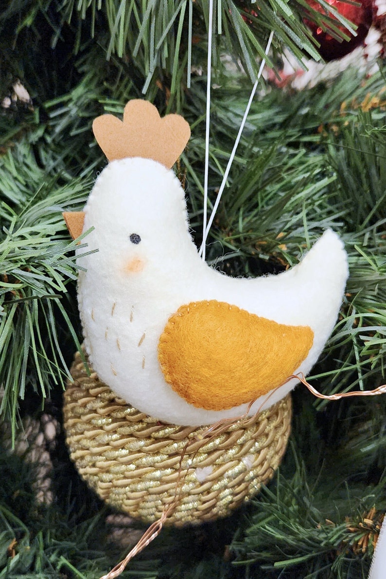 May include: A white felt bird ornament with a brown felt comb and a yellow felt spot on its chest. The bird is sitting in a gold glitter nest.