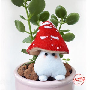 May include: A plush mushroom toy with a red and white cap and a white body. The toy is sitting in a pink pot with brown stones.