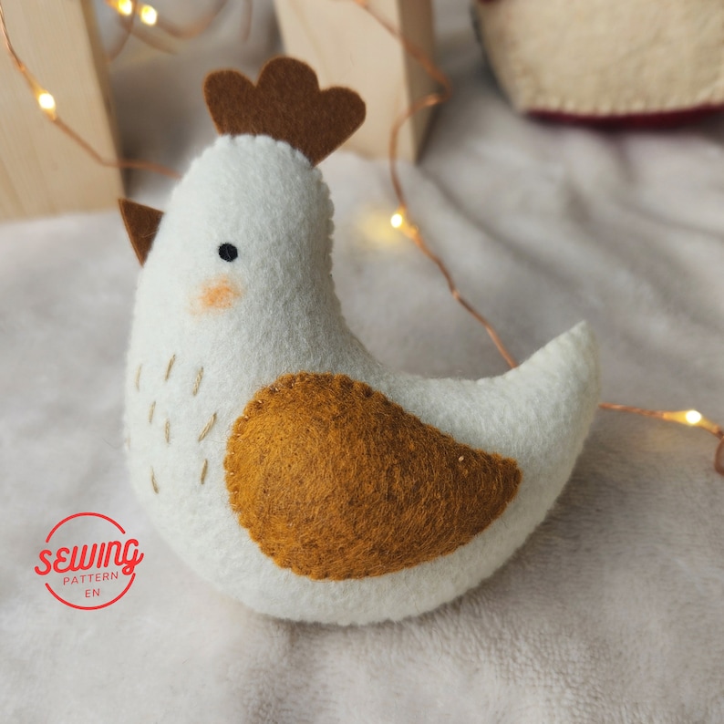 May include: A white felt chicken with a brown felt comb and wattle. The chicken has a brown felt patch on its back. The chicken is sitting on a white fluffy surface.