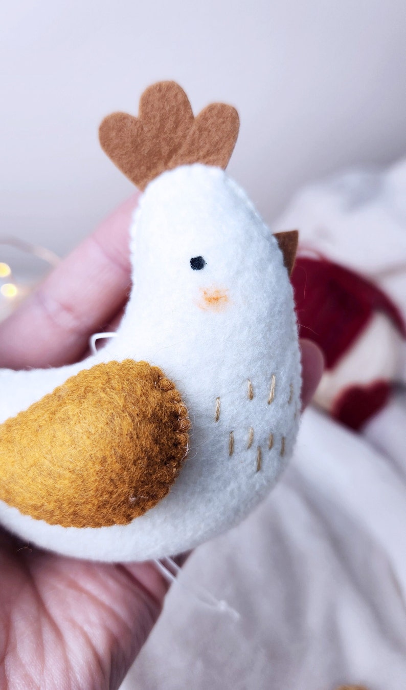 May include: A white felt chicken ornament with a brown felt comb and a yellow felt wing. The chicken has embroidered details and a stitched beak.