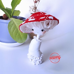 Könnte beinhalten: Eine weiße Stoffpuppe mit einem rot-weißen Pilz-Hut. Die Puppe trägt ein weißes Kleid mit Rüschen und hat einen weißen pilzförmigen Sockel. Die Puppe steht auf einer weißen Oberfläche. "Sewing Pattern EN" steht in Rot auf einem weißen Kreis.