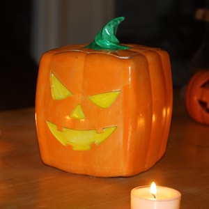May include: A ceramic jack-o'-lantern with a carved face and a green stem. The pumpkin is lit from inside, creating a warm glow.