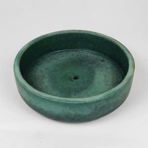 Può includere: Un vaso rotondo in ceramica verde con una superficie leggermente ruvida. Il vaso ha un piccolo foro di drenaggio al centro.