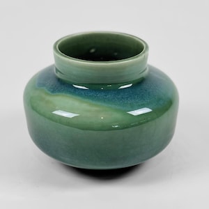 Puede incluir: Un jarrón de cerámica pequeño y rechoncho con un cuerpo ancho y redondeado y un cuello corto y erguido. La superficie presenta un esmalte brillante y suave en varios tonos de verde salvia y azul cerceta, creando un aspecto fluido y mezclado.
