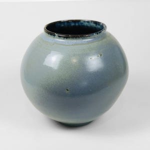 Puede incluir: Un jarrón redondo de cerámica azul claro con un borde azul oscuro. El jarrón tiene un acabado liso y brillante.
