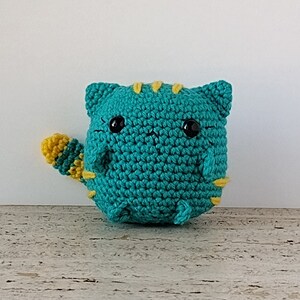 May include: A teal crocheted cat with yellow stripes and a yellow tail. The cat has black eyes and a cute expression.