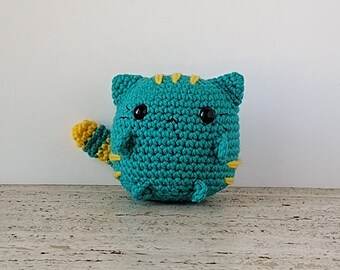 May include: A teal crocheted cat with yellow stripes and a yellow tail. The cat has black eyes and a cute expression.