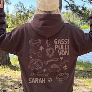 Könnte beinhalten: Ein dunkelbrauner Kapuzenpullover mit einer cremefarbenen Grafik auf der Rückseite. Das Design zeigt Illustrationen von Hundeaccessoires wie Halsband, Leine, Geschirr, Napf und Knochen sowie den Text GASSI PULLI VON SARAH und Pfotenabdrücke.