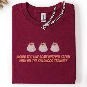 Könnte beinhalten: Burgunderfarbenes T-Shirt mit dem Text "WOULD YOU LIKE SOME WHIPPED CREAM WITH ALL THE CHILDHOOD TRAUMA?" und drei Sahne-Grafiken mit Erdbeeren. Eine silberne Halskette liegt über dem Shirt.