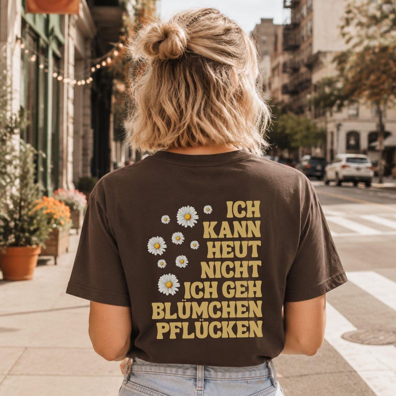 May include: A dark brown short-sleeved t-shirt featuring a graphic on the back with white daisy illustrations and the yellow text "ICH KANN HEUT NICHT ICH GEH BL&Uuml;MCHEN PFL&Uuml;CKEN" arranged in a block layout on the right side of the shirt.