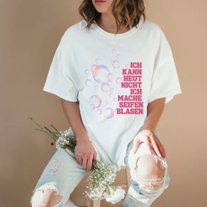 May include: A white short-sleeved t-shirt featuring a graphic of iridescent soap bubbles and the pink text "ICH KANN HEUT NICHT ICH MACHE SEIFENBLASEN" printed on the front. The shirt has a crew neckline and a relaxed fit, showcasing a casual style.