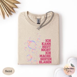 May include: A sand-colored t-shirt featuring a graphic of iridescent bubbles and the pink text "ICH KANN HEUT NICHT ICH MACHE SEIFENBLASEN". A silver-toned metal chain is attached to the neckline of the shirt, which has a standard crew neck design.