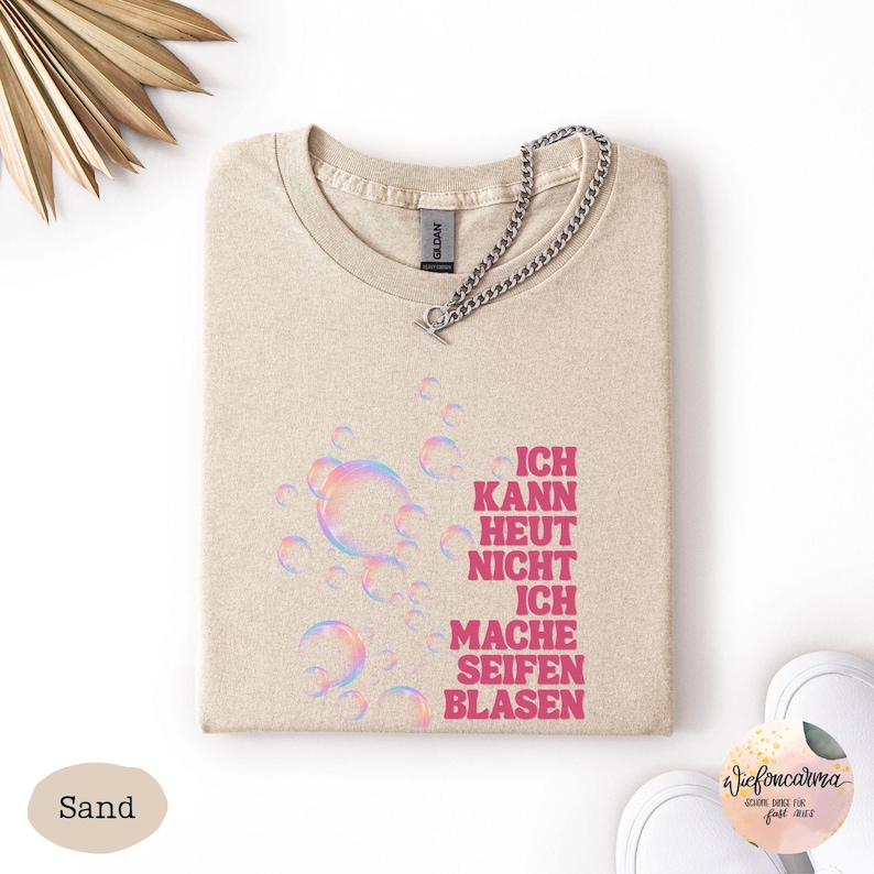 May include: A sand-colored t-shirt featuring a graphic of iridescent bubbles and the pink text "ICH KANN HEUT NICHT ICH MACHE SEIFENBLASEN". A silver-toned metal chain is attached to the neckline of the shirt, which has a standard crew neck design.