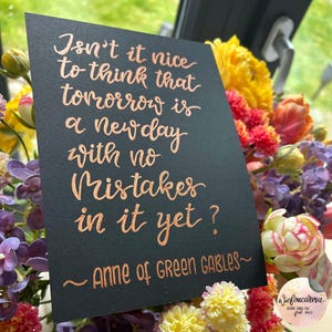 Könnte beinhalten: Eine schwarze Karte mit kupferfarbener Kalligraphie mit der Aufschrift "Isn't it nice to think that tomorrow is a new day with no mistakes in it yet?" und "Anne of Green Gables~". Die Karte ist von bunten Blumen umgeben.
