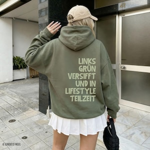 Könnte beinhalten: Ein olivgrüner Kapuzenpullover mit dem aufgedruckten Text LINKS GRÜN VERSIFFT UND IN LIFESTYLE TEILZEIT in hellgrüner Schrift auf der Rückseite, kombiniert mit einem weißen Faltenrock und einer beigen Baseballkappe.