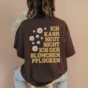 May include: A dark brown short-sleeve t-shirt featuring a graphic on the back with white daisy illustrations and the yellow text "ICH KANN HEUT NICHT ICH GEH BL&Uuml;MCHEN PFL&Uuml;CKEN" arranged in a block format on the right side of the garment.