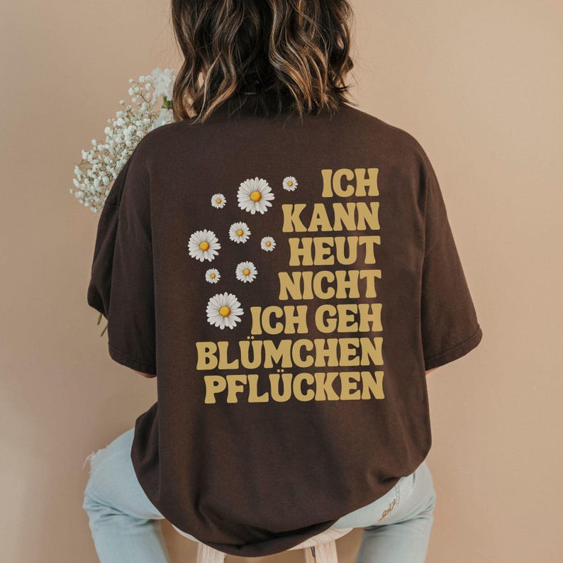 May include: A dark brown short-sleeve t-shirt featuring a graphic on the back with white daisy illustrations and the yellow text "ICH KANN HEUT NICHT ICH GEH BL&Uuml;MCHEN PFL&Uuml;CKEN" arranged in a block format on the right side of the garment.