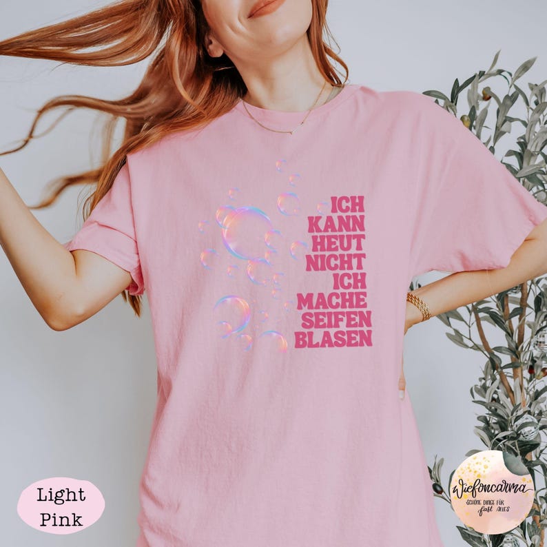 May include: A light pink short-sleeved t-shirt featuring a graphic of iridescent bubbles on the left side and the German text "ICH KANN HEUT NICHT ICH MACHE SEIFENBLASEN" printed in bold, dark pink capital letters on the right side of the chest.