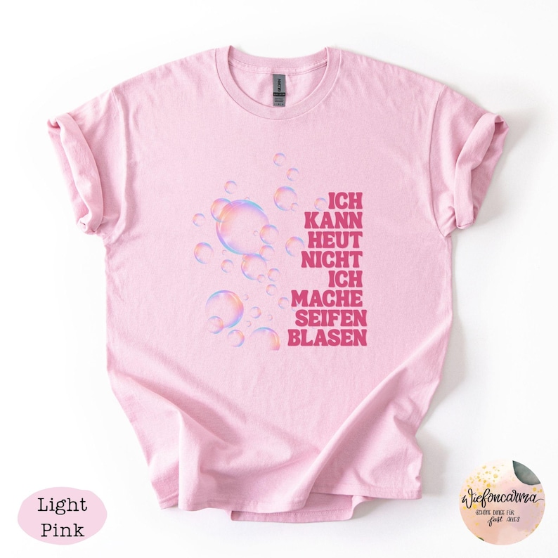 May include: A light pink t-shirt featuring a graphic of iridescent soap bubbles and the German text "ICH KANN HEUT NICHT ICH MACHE SEIFEN BLASEN" printed in dark pink. The shirt has short sleeves and a crew neckline, with a simple, casual design.