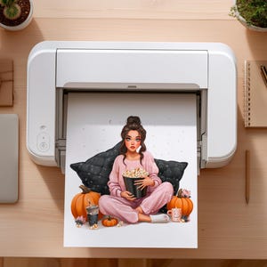 May include: A print of a woman in pink pajamas holding a popcorn bucket, sitting on a black pillow. Two orange pumpkins, a drink, and a mug are also in the image. The print is coming out of a white printer.