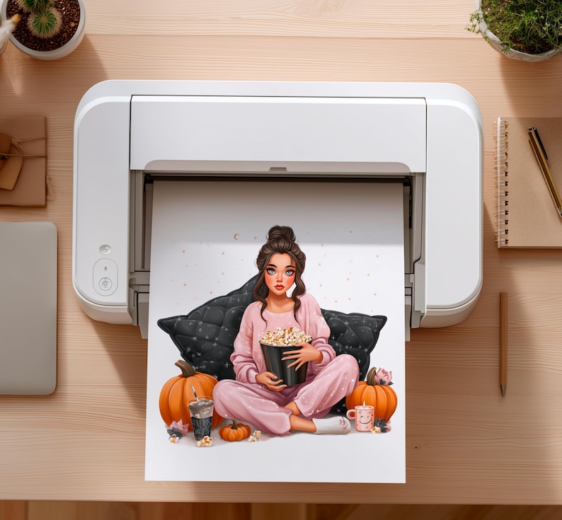 May include: A print of a woman in pink pajamas holding a popcorn bucket, sitting on a black pillow. Two orange pumpkins, a drink, and a mug are also in the image. The print is coming out of a white printer.