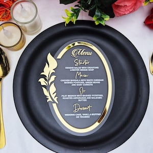 May include: A black plate with a gold-rimmed menu card featuring a gold floral design. The menu card lists a starter, main course, and dessert options. Gold utensils and candles are also present.