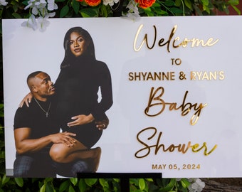 May include: A white sign with gold lettering announcing a baby shower for Shyanne & Ryan on May 05, 2024. A photo of the couple is featured on the sign.