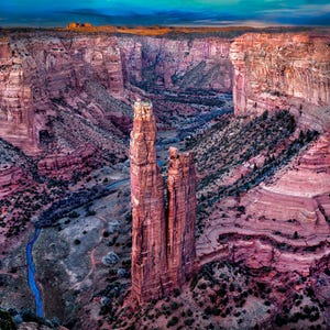 May include: A tall, thin rock formation rises from a canyon floor, with a blue stream winding through the red rock landscape. The rock formation is a striking example of natural erosion.