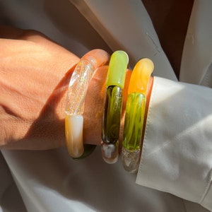 Pu&ograve; includere: Tre braccialetti di vetro colorati impilati su un polso. I braccialetti sono traslucidi e presentano una variet&agrave; di colori, tra cui verde, giallo e bianco.