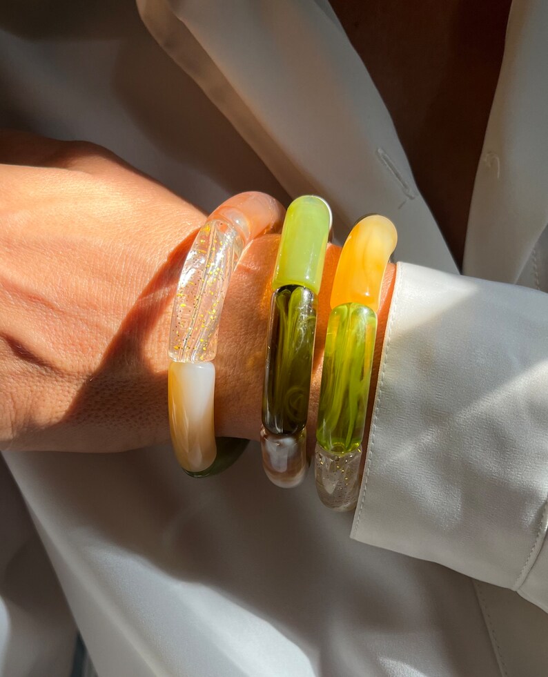 Pu&ograve; includere: Tre braccialetti di vetro colorati impilati su un polso. I braccialetti sono traslucidi e presentano una variet&agrave; di colori, tra cui verde, giallo e bianco.