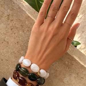 May include: Three beaded bracelets in shades of white, green, and brown. The bracelets are made of large, oval-shaped beads. A gold initial charm is visible on one bracelet. The person is wearing gold rings. The person is wearing a dark green blazer.