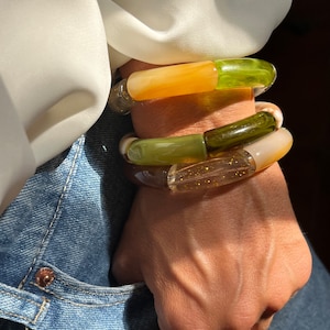 May include: Three stacked bracelets made of green, brown, and orange resin. The bracelets are layered on a person's wrist.