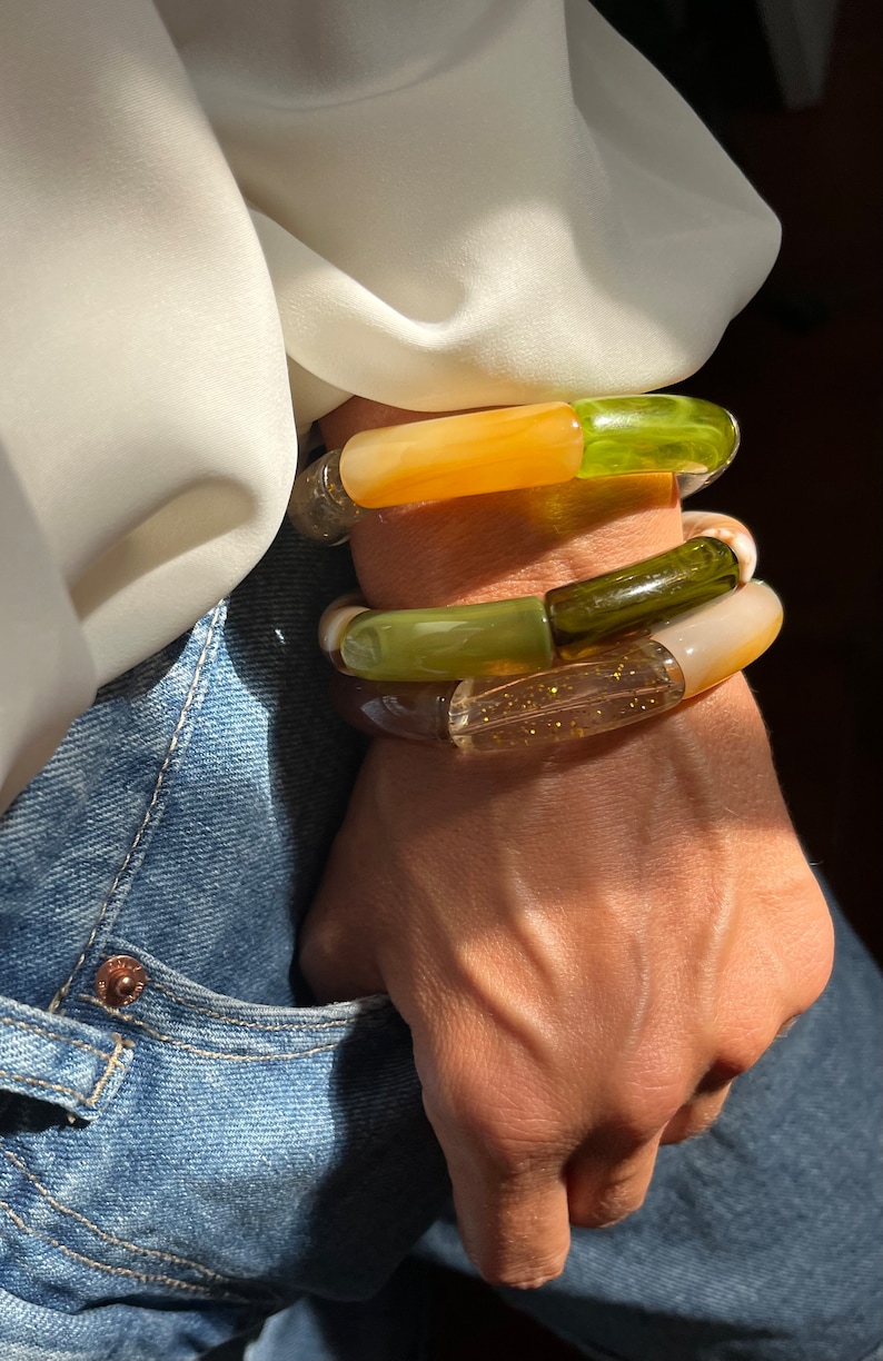 Pu&ograve; includere: Tre braccialetti impilati in resina verde, marrone e arancione. I braccialetti sono sovrapposti al polso di una persona.