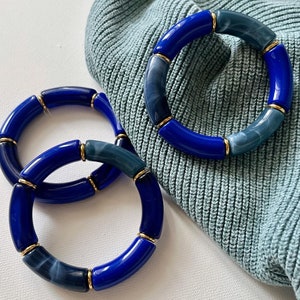 Könnte beinhalten: Drei blaue und goldfarbene Armbänder. Die Armbänder bestehen aus Segmenten in verschiedenen Blautönen mit goldfarbenen Verbindern. Die Armbänder liegen auf einem hellblauen Strickstoff.
