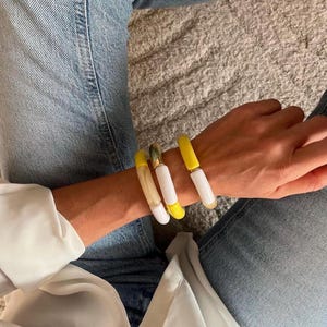 May include: Three yellow and white bangle bracelets with gold accents. The bracelets are stacked on a person's wrist.